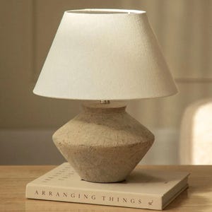 May include: A small, beige table lamp with a white lampshade. The lamp has a textured, vase-shaped base and sits on a closed book with the title "ARRANGING THINGS". The lamp is designed for indoor use and provides soft lighting.