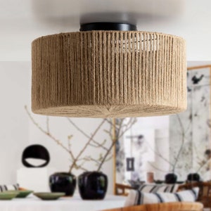 May include: A round, woven, natural fibre ceiling light fixture with a black metal base. The light fixture is hanging from a ceiling in a room with a white wall and a wooden floor.