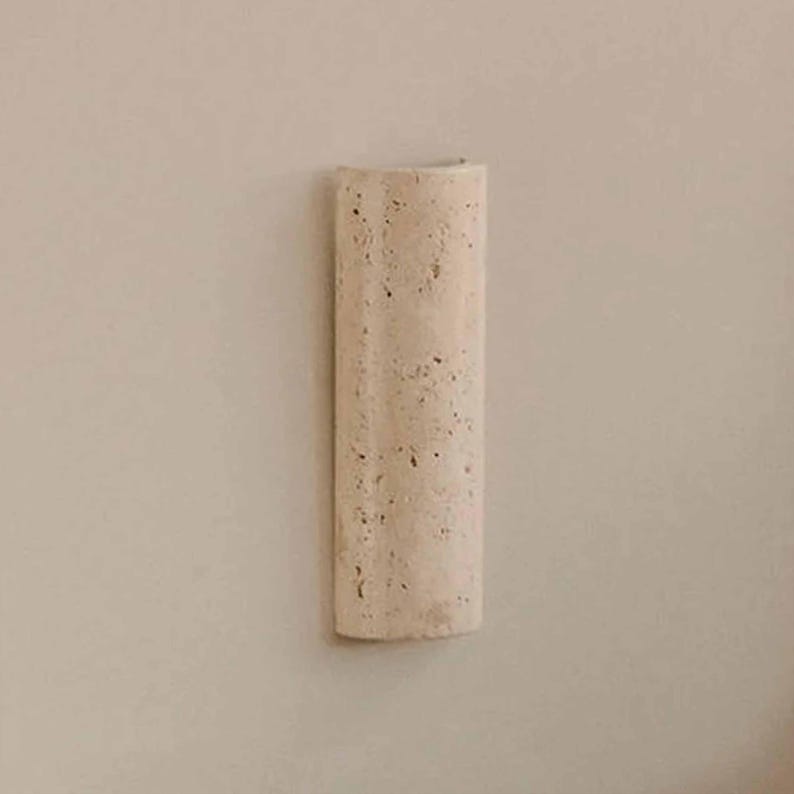 May include: A beige, rectangular wall sconce with a curved top. The sconce has a textured surface with small, dark spots. The design is minimalist and modern, suitable for interior decor.