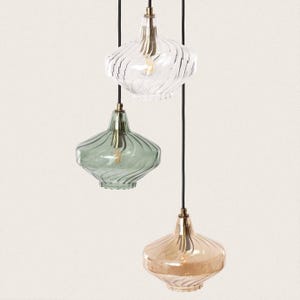 Peut inclure: Trois lampes suspendues en verre avec un motif en spirale. Les lampes sont suspendues à des cordons noirs et ont une finition en laiton. La lampe du haut est en verre clair, la lampe du milieu est en verre vert et la lampe du bas est en verre ambre.