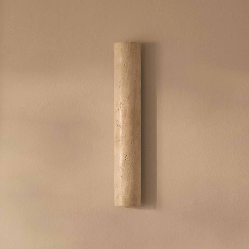 May include: A cylindrical, beige-colored wall sconce with a textured surface. The sconce is vertically oriented and appears to be made of stone or a similar material. The background is a solid, neutral color.