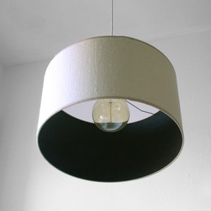 May include: A pendant light fixture with a white lampshade and a dark interior. The light bulb is visible through the opening. The fixture hangs from a white cord against a white wall.