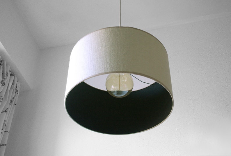 May include: A pendant light fixture with a white lampshade and a dark interior. The light bulb is visible through the opening. The fixture hangs from a white cord against a white wall.
