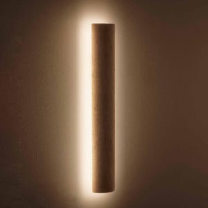 May include: A cylindrical wall sconce made of light brown cork, illuminated by a vertical strip of warm white light. The sconce is mounted on a textured, neutral-toned wall, creating a minimalist and modern aesthetic.