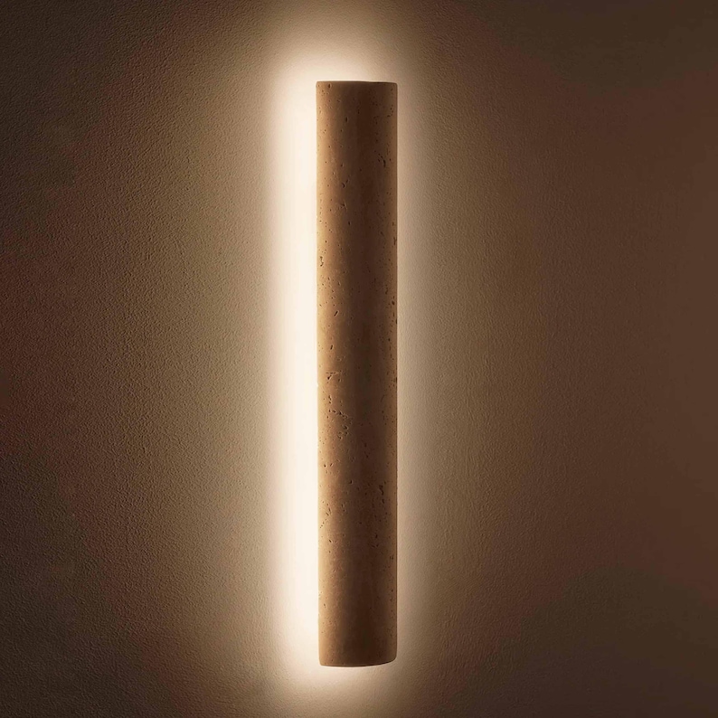 May include: A cylindrical wall sconce made of light brown cork, illuminated by a vertical strip of warm white light. The sconce is mounted on a textured, neutral-toned wall, creating a minimalist and modern aesthetic.