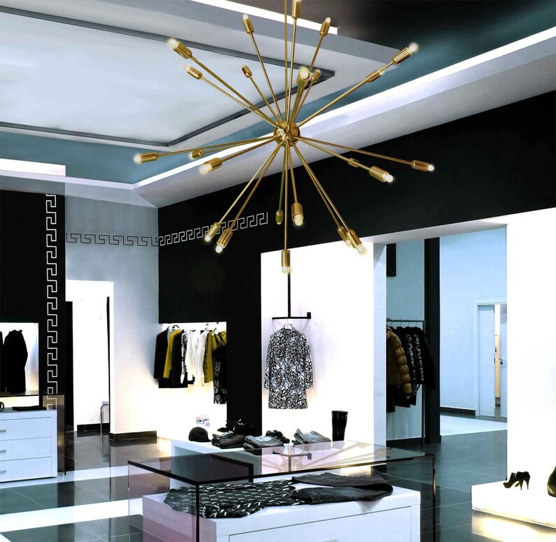May include: A gold-toned, multi-arm chandelier with multiple light bulbs hangs from the ceiling of a modern retail store. The chandelier has a starburst design with long, thin arms extending outward from a central hub.