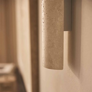 May include: A close-up of a beige, cylindrical, textured wall sconce. The sconce is made of a porous material and is mounted on a light-colored wall. The design is minimalist and modern.