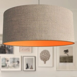 May include: A cylindrical lampshade with a textured, light gray exterior and a bright orange interior. The lampshade is suspended from the ceiling, and the background features framed artwork.