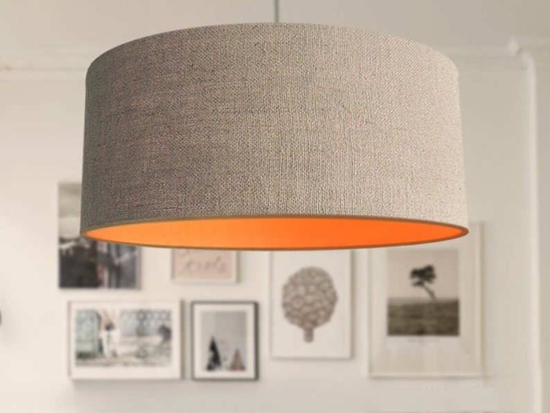 May include: A cylindrical lampshade with a textured, light gray exterior and a bright orange interior. The lampshade is suspended from the ceiling, and the background features framed artwork.