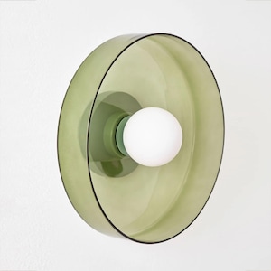 May include: A modern, wall-mounted light fixture with a pale green glass shade and a white, spherical bulb. The shade has a circular design with a black trim. The fixture is mounted on a white wall, creating a minimalist aesthetic.