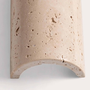 May include: A section of a light beige, curved stone architectural element. The stone has a porous texture with small holes and imperfections. The bottom edge is a rounded arch, and the piece is set against a white background.