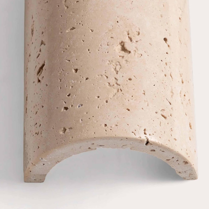 May include: A section of a light beige, curved stone architectural element. The stone has a porous texture with small holes and imperfections. The bottom edge is a rounded arch, and the piece is set against a white background.