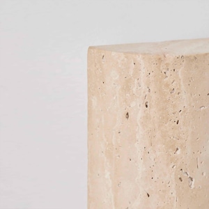 May include: A close-up of a beige, rectangular stone block with a rough, textured surface. The stone has small holes and imperfections, and the edges are slightly rounded. The background is plain white.