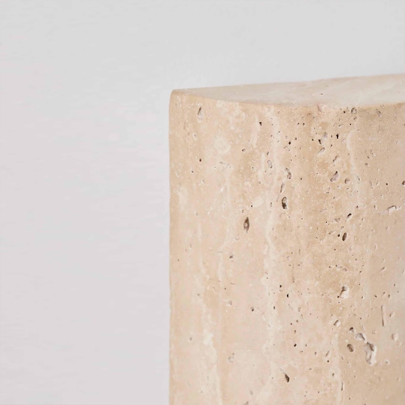May include: A close-up of a beige, rectangular stone block with a rough, textured surface. The stone has small holes and imperfections, and the edges are slightly rounded. The background is plain white.