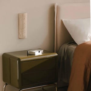 May include: A modern olive green nightstand with two drawers and a silver box on top. A beige, textured wall sconce is mounted above the nightstand. A bed with a white pillow and brown blanket is in the background.