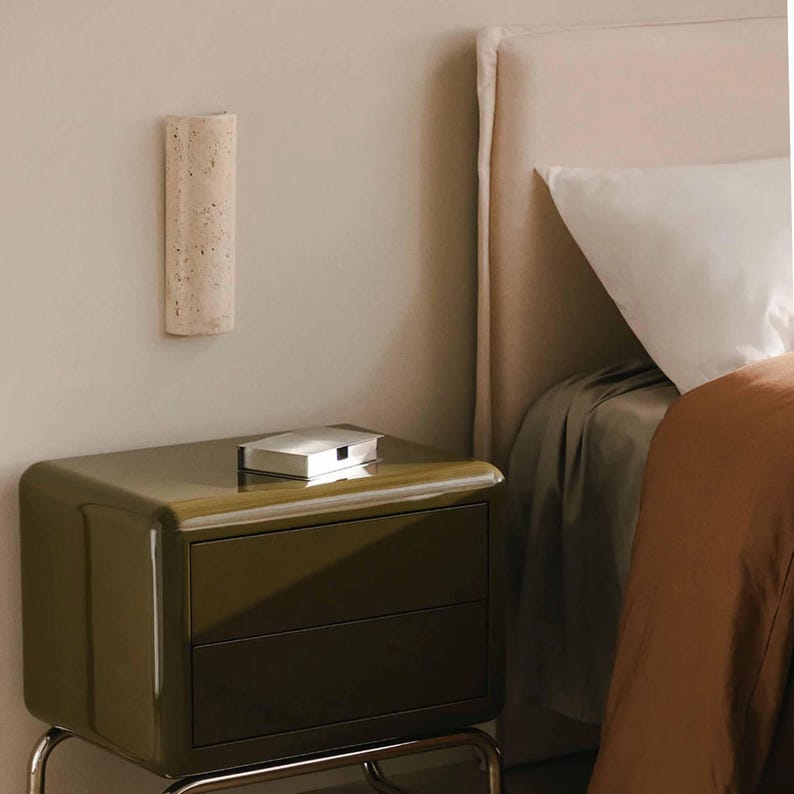 May include: A modern olive green nightstand with two drawers and a silver box on top. A beige, textured wall sconce is mounted above the nightstand. A bed with a white pillow and brown blanket is in the background.