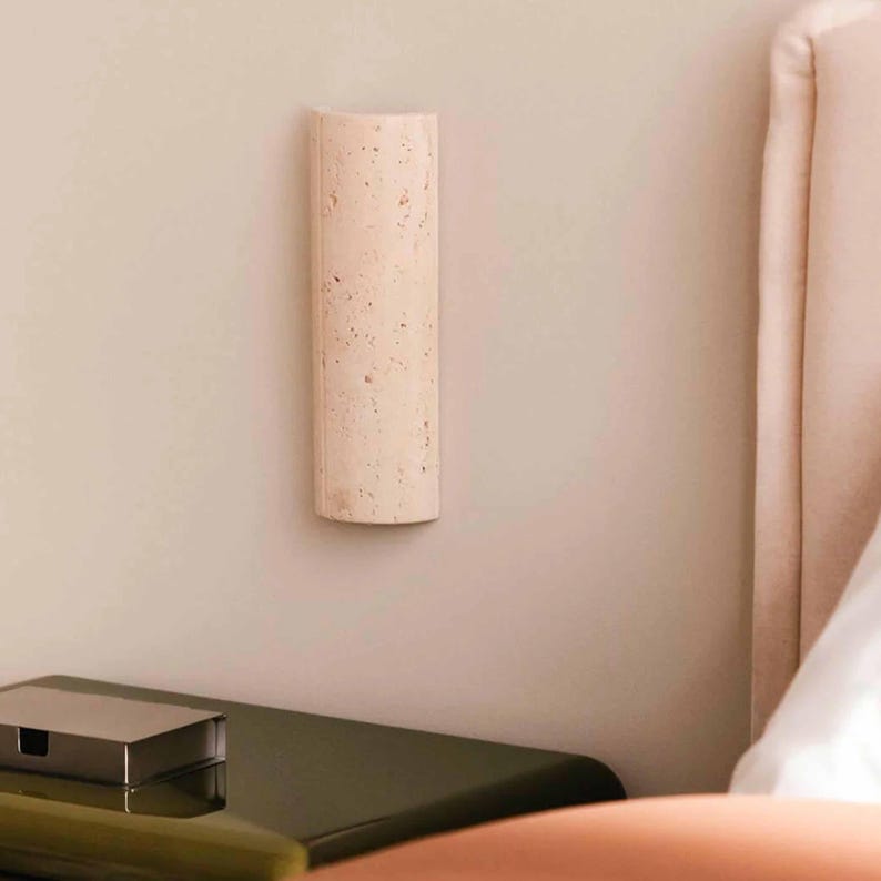 May include: A light beige, rectangular wall sconce with a rounded top is mounted on a light pink wall. The sconce has a textured surface with small, dark spots. A dark green nightstand and a bed with a pink headboard are in the background.