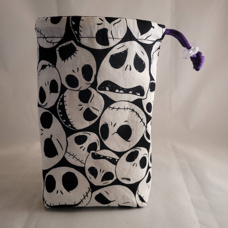 May include: A black and white fabric dice bag with a repeating pattern of Jack Skellington's face from the Nightmare Before Christmas. The bag has a purple drawstring with a clear plastic toggle.