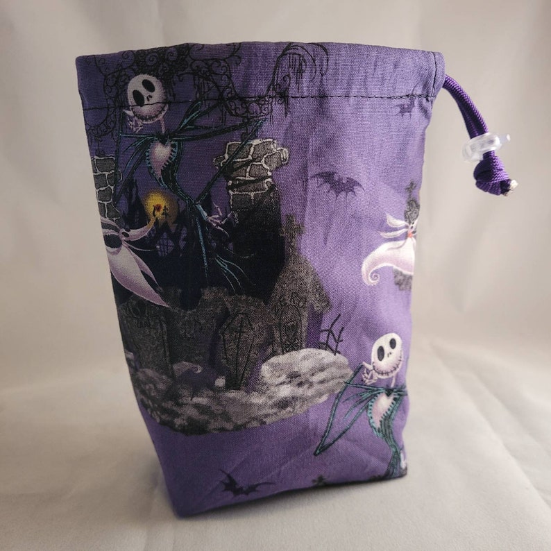 May include: Purple fabric drawstring bag with a repeating pattern of Jack Skellington and Sally from The Nightmare Before Christmas. The bag has a white drawstring.