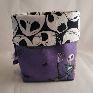May include: A purple fabric storage bin with a black and white Nightmare Before Christmas print. The bin has a white fabric lining with a black and white skull pattern.