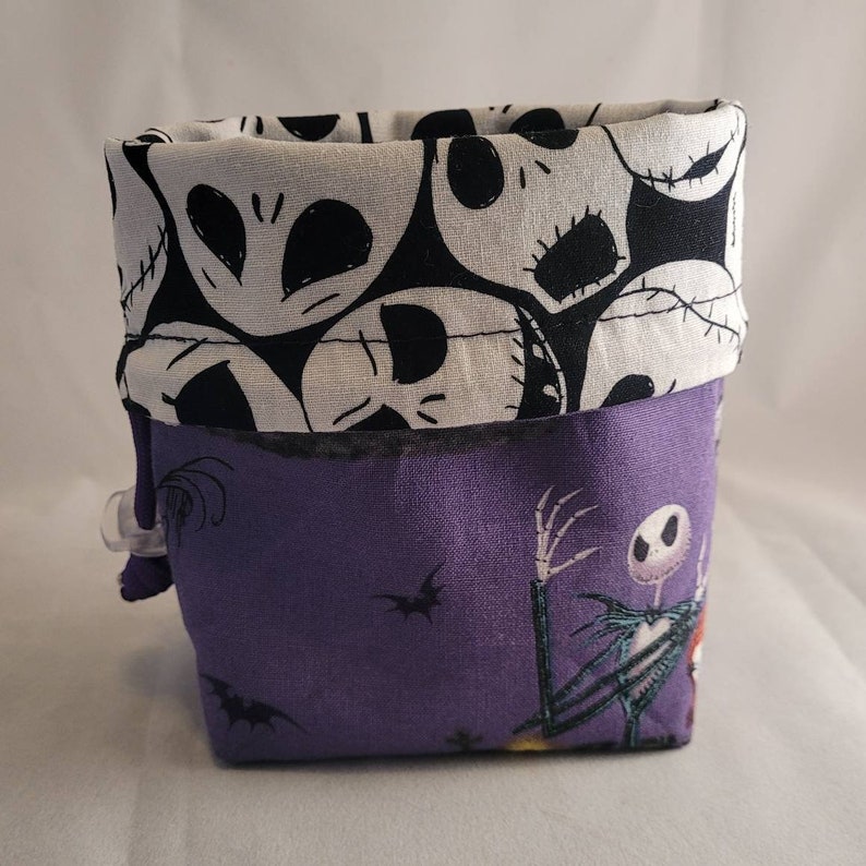 May include: A purple fabric storage bin with a black and white Nightmare Before Christmas print. The bin has a white fabric lining with a black and white skull pattern.
