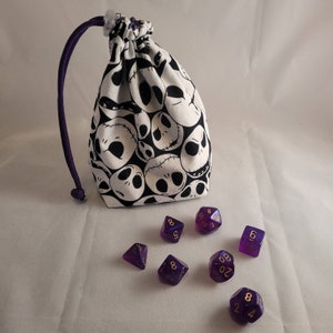 May include: A white fabric dice bag with a black and white pattern of cartoon skulls. The bag is open and there are eight purple dice with gold numbers on a white surface.