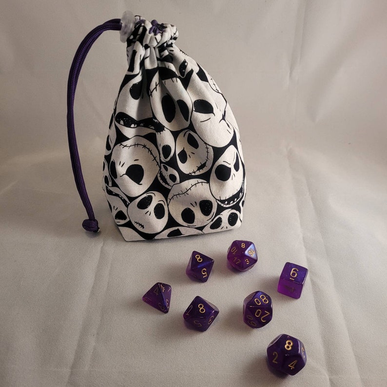 May include: A white fabric dice bag with a black and white pattern of cartoon skulls. The bag is open and there are eight purple dice with gold numbers on a white surface.