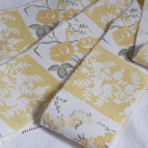 Puede incluir: Primer plano de varios trozos de tela con estampado floral. La tela presenta un diseño repetido de rosas amarillas y follaje blanco sobre un fondo blanco. Los bordes de la tela están deshilachados y las piezas están dispuestas sobre una superficie blanca.