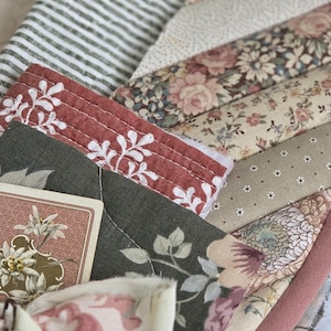 May include: A quilted fabric piece featuring a patchwork of floral, striped, and solid patterns in muted tones of dusty rose, olive green, cream, and charcoal. The item includes a tied fabric bow and a small rectangular card with an edelweiss illustration.
