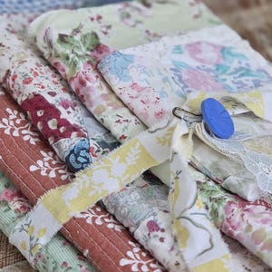 May include: A stack of folded fabric pieces featuring various floral patterns in shades of red, green, yellow, and blue. The fabric is secured with a fabric tie and a single blue button, and includes a decorative piece of white lace trim.
