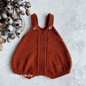 Könnte beinhalten: Ein gestrickter Baby-Strampler in Rostorange mit einem zarten Spitzenmuster. Der Strampler hat Träger und Holzknöpfe.