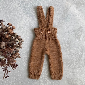 Op de afbeelding: Bruine gebreide baby-overall met bretels en twee bruine knopen.
