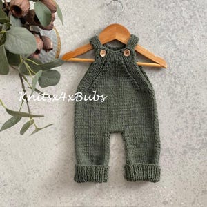 May include: Olive green knitted baby overalls with wooden buttons, displayed on a wooden hanger. The overalls have a ribbed cuff at the ankles. The text "Knits4xBubs" is visible in the image.