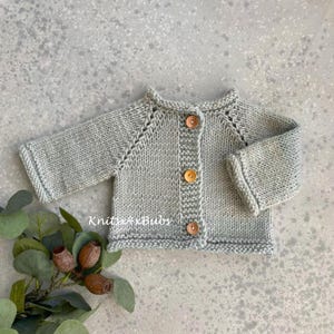 Könnte beinhalten: Hellgraue gestrickte Baby-Strickjacke mit drei Holzknöpfen. Die Strickjacke hat lange Ärmel und einen runden Halsausschnitt. Das Kleidungsstück ist aus weichem, strukturiertem Garn gefertigt. Ein handgefertigter Artikel, ideal für ein Neugeborenes.