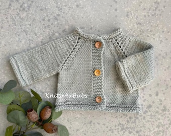May include: A light gray knitted baby cardigan with three wooden buttons. The cardigan has long sleeves and a round neckline. The garment is made of a soft, textured yarn, perfect for a newborn. The cardigan is a handmade item.