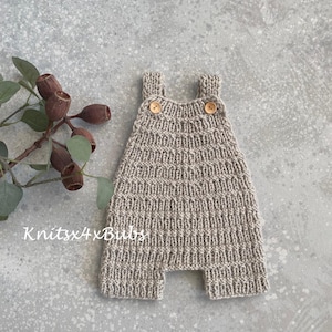 Può includere: Un pagliaccetto per neonati lavorato a maglia di colore beige neutro. Il pagliaccetto presenta un motivo a punto strutturato, bottoni in legno sulle spalline e il testo "Knitsx4xBubs".