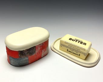 May include: A ceramic butter dish with a red, black, and blue geometric pattern. The lid is white and the dish has a matching white base. The dish is filled with a stick of butter labeled "Grade AA Butter".