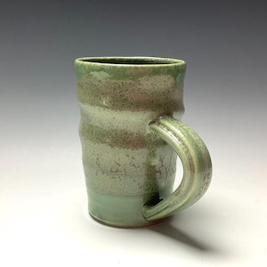 May include: A handmade ceramic mug with a light green glaze. The mug has a cylindrical shape with a rounded handle. The glaze has a textured appearance, with variations in color and tone. The mug is suitable for hot or cold beverages.