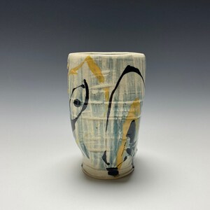 May include: A ceramic cup with a white glaze and abstract black and yellow painted designs.