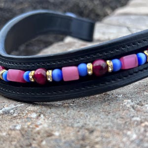 Könnte beinhalten: Schwarzes Leder-Stirnband mit dekorativem Perlendesign. Die Perlen sind eine Mischung aus Burgunderrot, Rosa, Blau und Gold. Das Stirnband ist gebogen und scheint für ein Pferd zu sein.