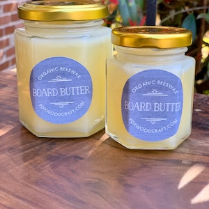 May include: Two glass jars with gold lids filled with a yellow substance. The jars have labels that say "Organic Beeswax Board Butter" and "PDXWOODCRAFT.COM".