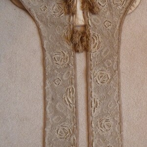 May include: A beige lace shawl with a scalloped edge and tassel trim. The shawl is made of delicate lace with a floral pattern.