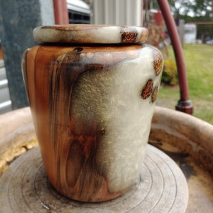 May include: A wooden urn with a natural wood grain pattern and a clear epoxy resin finish. The urn has a light brown, dark brown, and white color scheme. The urn is sitting on a pottery wheel.