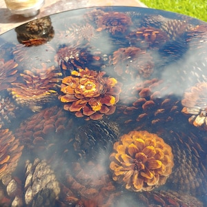 Może przedstawiać: Okrągły blat stołu z przezroczystym, błyszczącym wykończeniem, osadzony licznymi szyszkami. Szyszki, o różnych rozmiarach, mają głównie odcienie brązu i pomarańczy, tworząc naturalny, teksturowany wzór. Średnica: około 76 cm.