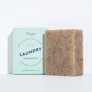 Op de afbeelding: Een lichtblauwe doos met de tekst "Tangie Laundry Concentrate Eco Friendly Refillable Liquid Laundry Soap wastefreeproducts.com" en een reep bruine waszeep.