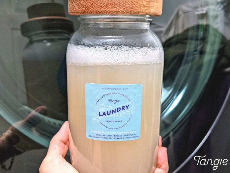Op de afbeelding: Een glazen pot gevuld met lichtbruine vloeibare wasmiddelzeep. De pot heeft een houten deksel en een blauw en wit etiket met de tekst "Tangle Laundry Liquid Soap". Het etiket bevat ook gebruiksaanwijzingen, zoals de hoeveelheid zeep die moet worden gebruikt voor verschillende wasladingen. De pot staat voor een wasmachine.