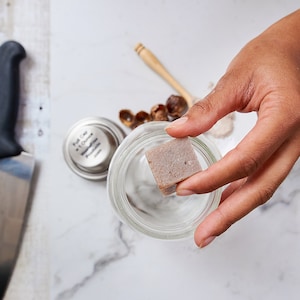 Op de afbeelding: Een hand houdt een kleine, vierkante, lichtbruine zeepstaaf boven een glazen pot. De zeepstaaf is licht getextureerd. De pot staat op een wit marmeren oppervlak.