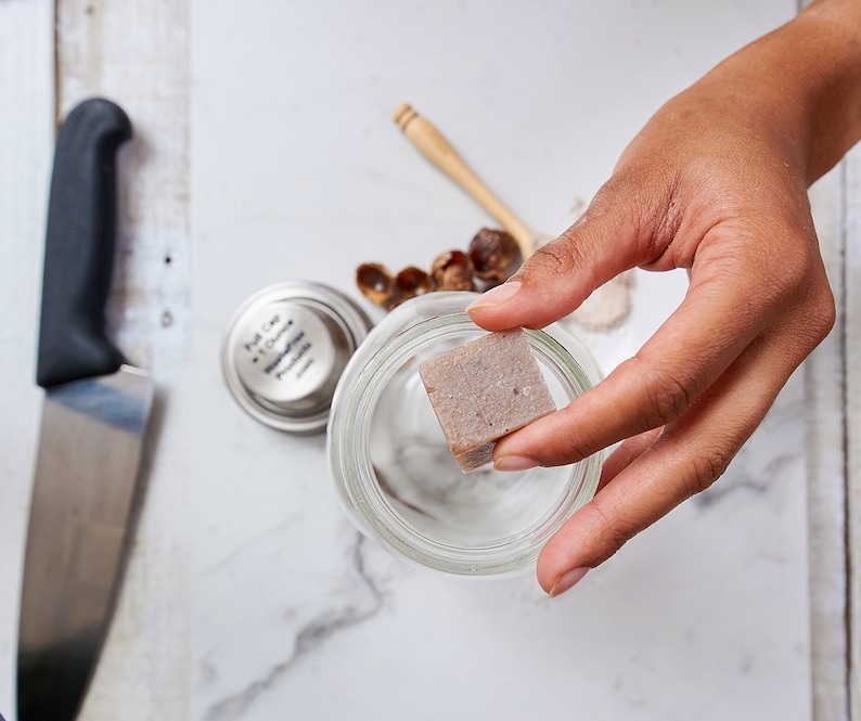 Op de afbeelding: Een hand houdt een kleine, vierkante, lichtbruine zeepstaaf boven een glazen pot. De zeepstaaf is licht getextureerd. De pot staat op een wit marmeren oppervlak.