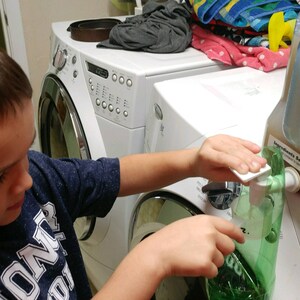 Op de afbeelding: Een jong persoon giet wasmiddel uit een grote plastic kan in een groene plastic zak. De kan heeft een etiket met de tekst "Keep Cap Loose".