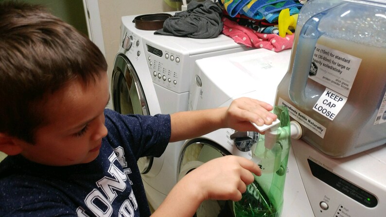 Op de afbeelding: Een jong persoon giet wasmiddel uit een grote plastic kan in een groene plastic zak. De kan heeft een etiket met de tekst "Keep Cap Loose".